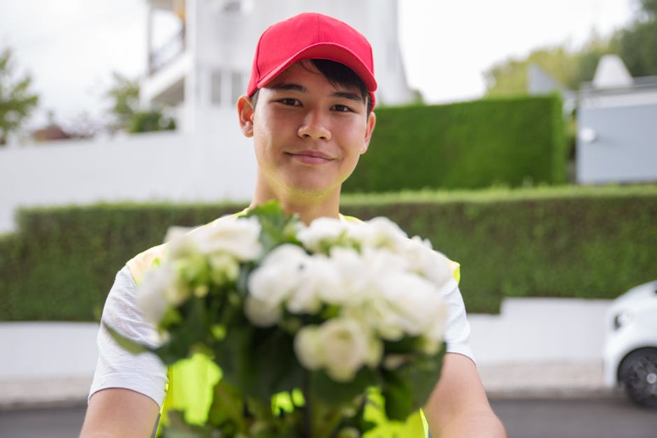 A delivery person with a friendly expression, wearing a red cap and a high-visibility jacket, holding a bouquet of fresh white flowers with green stems and leaves. The flowers are arranged in a rounde