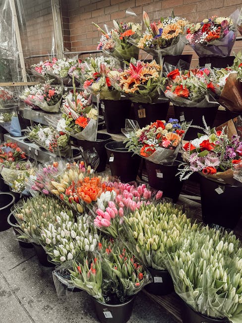 A wide selection of fresh floral bouquets and individual flower arrangements displayed in black buckets against a brick wall. The flowers include pink, white, and red tulips, orange roses, and mixed s