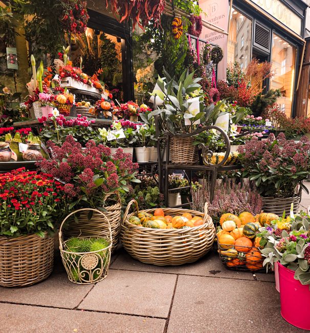 An outdoor flower display in front of a shop, featuring an assortment of fresh flowers and plants arranged in woven baskets and pots. Prominent are pink and purple chrysanthemums, white calla lilies,