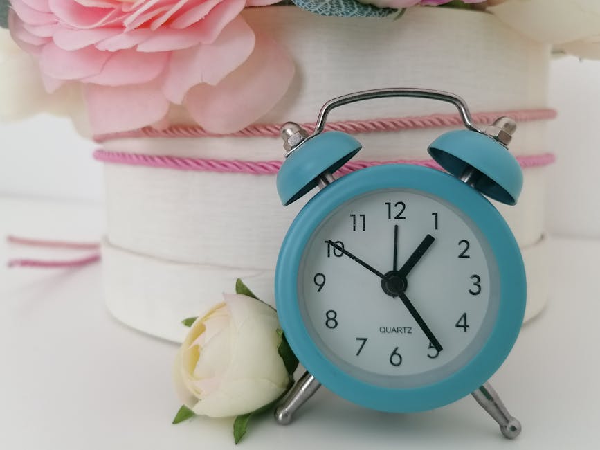 A light blue vintage-style alarm clock showing the time as 12:05, placed on a white surface with a small white rose with green leaves leaning against its base. In the background, there is a round, whi