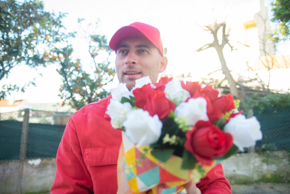 A delivery person in a red uniform and matching cap holds a colorful floral bouquet, featuring red and white roses, with lush green foliage. The bouquet is wrapped in decorative paper with geometric p
