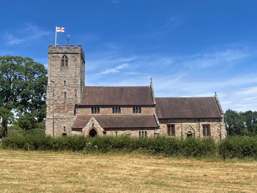 A historic stone church with a tall square tower topped by a flagpole flying the Union Jack, set against a bright blue sky with wispy clouds. The church features small arched windows and a pitched roo